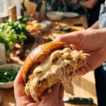 A close-up of two hands holding and slightly squeezing a messy shredded garlic parmesan chicken sandwich, with creamy sauce dripping out.