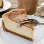 A close-up of a slice of snickerdoodle cheesecake showing a fork taking a bite from the creamy filling and thick cookie crust.