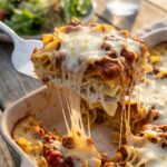 A close-up of a spatula lifting a generous, cheesy piece of beef noodle casserole out of the pan, showing layers of creamy noodles, meat sauce, and a long cheese pull.