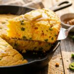 A close-up of a warm wedge of jalapeño cheddar cornbread being lifted from a skillet, with butter melting on top and a fluffy interior crumb.