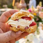A close-up of a hand holding a strawberry cream puff with a bite taken out, revealing the hollow pastry shell filled with cream and fruit.