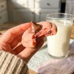 A close-up of a hand holding a pink strawberry cookie with a chocolate kiss, with a bite taken out showing the soft texture.