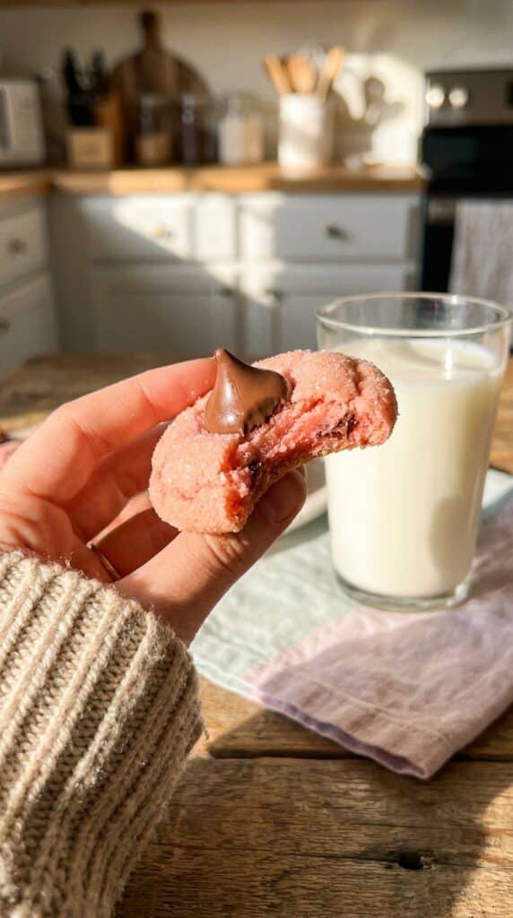 A close-up of a hand holding a pink strawberry cookie with a chocolate kiss, with a bite taken out showing the soft texture.