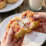 A close-up of hands pulling apart a warm pastry roll, revealing a gooey strawberry cheesecake center and a pink crumb topping.