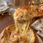 A close-up of a toasted piece of bread scooping hot, cheesy crab and shrimp dip out of a bread bowl, creating a stringy cheese pull.