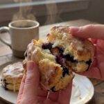 A close-up of hands pulling apart a warm blueberry biscuit, showing the flaky interior layers and juicy baked berries.