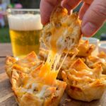 A close-up of a hand picking up a crispy chicken taco bite, creating a long, gooey cheese stretch from the serving board.