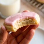 A close-up of a hand holding a pink frosted sugar cookie with a bite taken out, showing the soft, dense crumb inside.