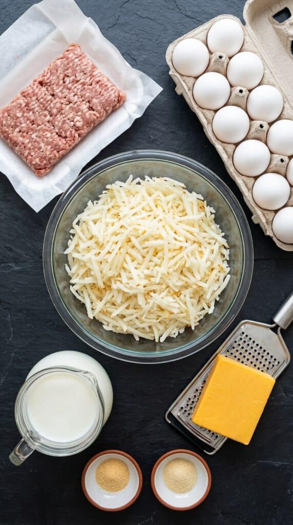 A flat lay showing hash browns, raw ground sausage, eggs, milk, a block of cheddar cheese, and spices on a dark slate board.