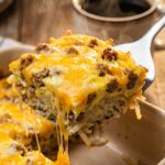 A close-up of a spatula lifting a hearty square slice of breakfast casserole, showing layers of hash browns, sausage, eggs, and stringy melted cheese.