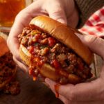 A close-up of two hands holding a messy sloppy joe sandwich, with the savory meat sauce dripping down the sides.