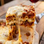 A close-up of a spatula lifting a thick, beautifully layered square of lasagna out of a pan, creating a massive stringy cheese pull.
