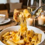 A close-up of a fork lifting a piece of chicken and pasta from a bowl, creating a thick, gooey stretch of melted cheddar cheese.