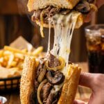 Two hands pulling apart a loaded steak and mushroom sandwich, revealing a massive, gooey cheese stretch, with fries in the background.