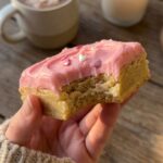 A close-up of a hand holding a frosted pink sugar cookie bar with a bite taken out, showing the soft, dense texture inside.