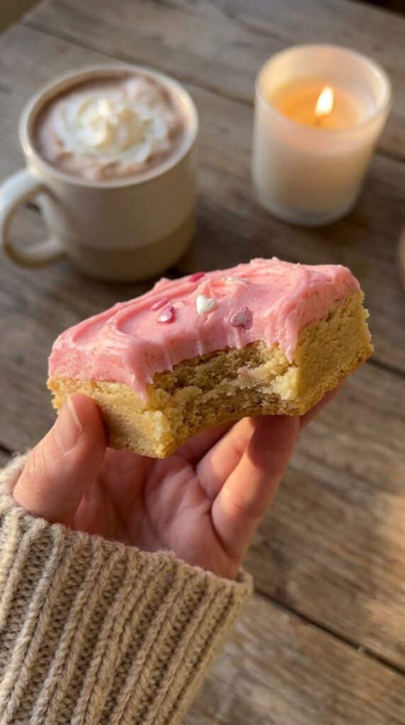 A close-up of a hand holding a frosted pink sugar cookie bar with a bite taken out, showing the soft, dense texture inside.