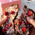 A close-up of two hands breaking a piece of decorated chocolate bark in half, with a pink gift box and champagne in the background.