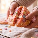 A close-up of hands breaking a soft, pink Valentine's cookie in half, showing the pillowy, cake-like center and chocolate chips.