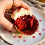 A close-up of a half-eaten red velvet cupcake with the wrapper being peeled back, showing the bright red, moist interior.