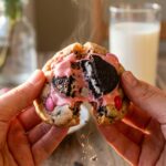 Two hands breaking a warm, soft cookie in half, revealing melted pink M&Ms and Oreo chunks inside.