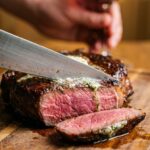 A close-up of a knife slicing a thick rib eye steak, revealing a juicy, pink medium-rare center with a dark, buttery crust.