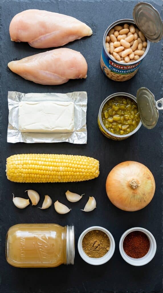 A flat lay showing raw chicken, white beans, cream cheese, green chiles, corn, chicken broth, and spices on a dark slate board.