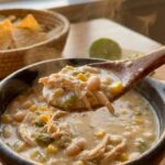 A close-up of a spoon lifting a thick, creamy bite of white chicken chili with beans and corn, with steam rising.