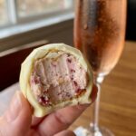 A close-up of a hand holding a white chocolate cheesecake ball with a bite taken out, revealing a creamy pink center with graham cracker and raspberry pieces.