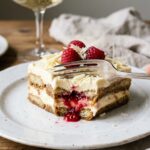 A close-up of a fork cutting into a slice of layered raspberry tiramisu, showing the creamy texture and juicy berries.