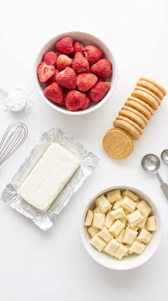 A flat lay showing freeze-dried strawberries, vanilla cookies, cream cheese, and white chocolate chunks on a white board.