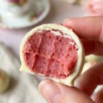 A close-up of a hand holding a white chocolate truffle with a bite taken out, revealing a dense pink strawberry center.