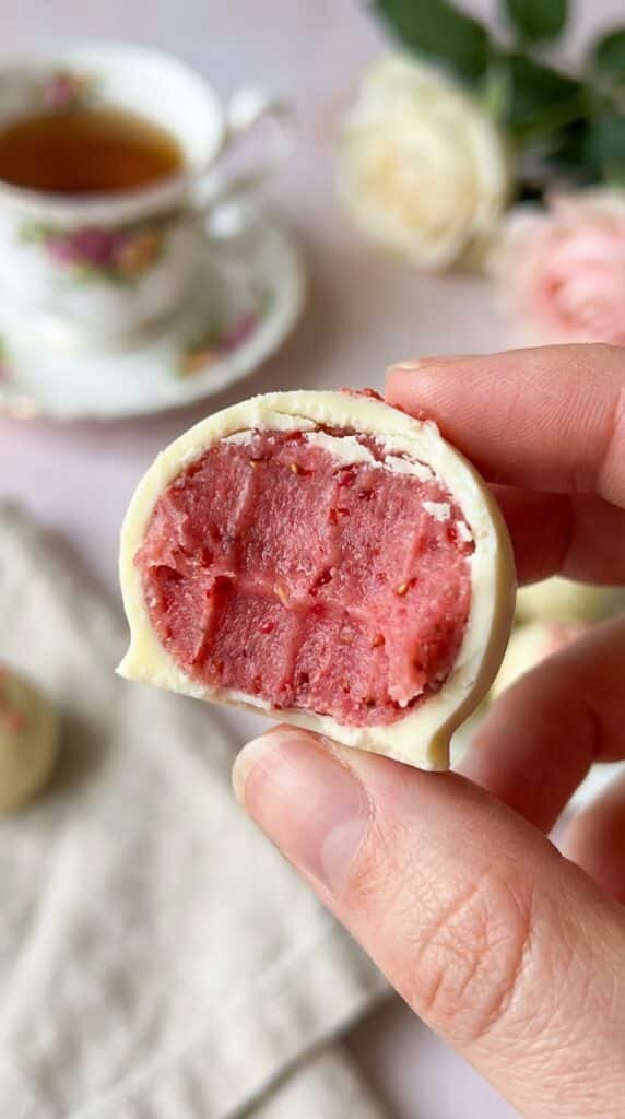A close-up of a hand holding a white chocolate truffle with a bite taken out, revealing a dense pink strawberry center.