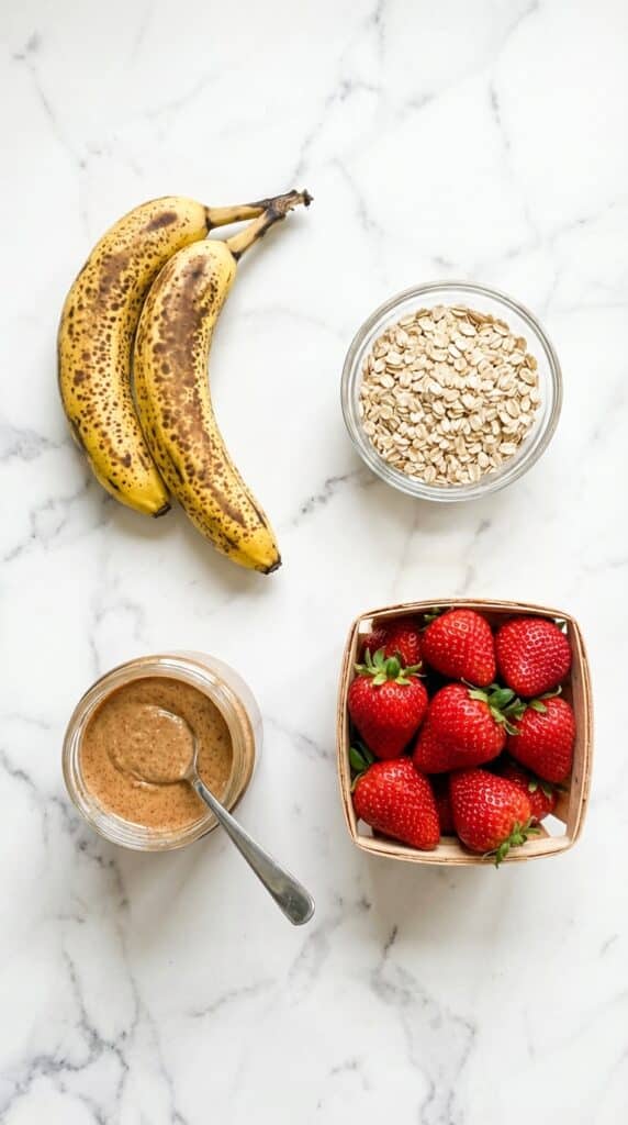 A flat lay showing two spotted bananas, a bowl of rolled oats, a jar of almond butter, and a basket of strawberries on a white marble counter.