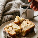 A close-up of a fork taking a bite of sliced apple fritter bread, highlighting the gooey cinnamon syrup and baked apples, with coffee in the background.