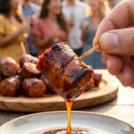 A close-up of a hand holding a toothpick with a glossy BBQ sausage bite, with a party scene blurred in the background.