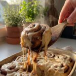 A close-up of a single gooey banana cinnamon roll being pulled from a baking pan, showing steam and melting icing