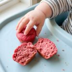 A close-up of a baby's hand reaching for a bright pink mini muffin on a blue highchair tray, with one muffin broken open to show the soft texture.