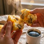 A close-up of hands breaking apart a warm savory sausage muffin, showing the steamy, cheesy interior.