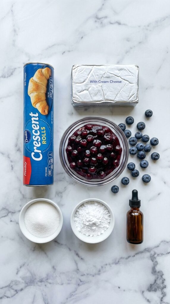A flat lay showing a tube of crescent dough, cream cheese, blueberry filling, powdered sugar, and vanilla on a marble board.