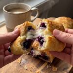 A close-up of hands pulling apart a warm blueberry cheesecake roll, showing the gooey cream cheese and blueberry filling inside