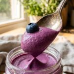 A close-up of a spoon lifting a thick, smooth bite of purple blueberry mousse with a fresh berry on top.