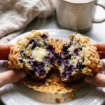 A close-up of a warm blueberry streusel muffin split in half with melting butter and bursting berries, next to a cup of coffee.