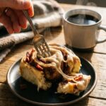 A close-up of a fork pulling apart the soft, steaming center of a bacon cinnamon roll covered in melting frosting.