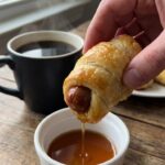 A close-up of a hand dipping a flaky sausage-stuffed crescent roll into a small bowl of maple syrup.