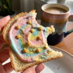 A close-up of a hand holding a piece of half-eaten rainbow unicorn toast with bright window light in the background.