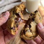 Two hands breaking apart a warm chocolate chip cookie, revealing a gooey melted chocolate pull, with a glass of milk in the background.