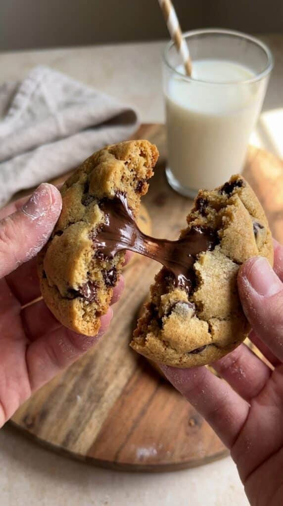 Two hands breaking apart a warm chocolate chip cookie, revealing a gooey melted chocolate pull, with a glass of milk in the background.