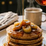 A close-up action shot of maple syrup being poured over a stack of banana pancakes with a coffee mug in the background.