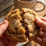 A close-up of hands pulling apart a warm, chewy brown sugar cookie, showing a soft, dense center topped with flaky sea salt.