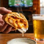 A close-up of a hand dipping a half-eaten buffalo chicken pastry bomb into a bowl of ranch dressing.